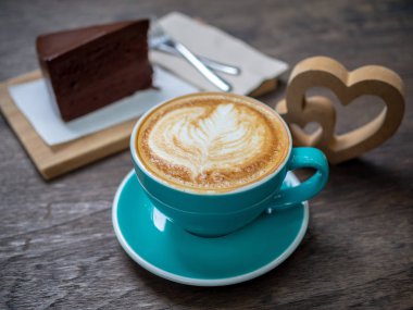 Kahve latte sanat bir parça çikolatalı kek, çatal ve ahşap masa arka plan üzerinde ahşap kalp ile mavi Kupası'nda. Kahve aşığı kavramı.