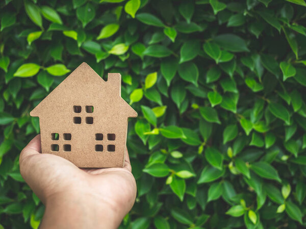 Wooden house on left hand on green leaves background with copy space. Home ecology concept.