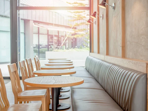 Ahşap sandalyeler, ahşap yuvarlak masalar, yerleşik deri kanepe ve lambalar ile ağaç arka plan üzerinde dışında ışıktan kafede cam pencere duvarı yakınındaki beton duvara satır.