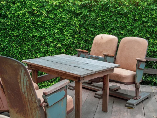 Vintage theatre seats with wooden table on wooden terrace on green bush background with copy space. Unique mix and match furniture style concept.