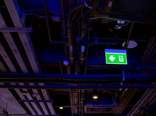 Illuminated green fire exit LED light sign on the ceiling in the dark with pipe line system inside the building.