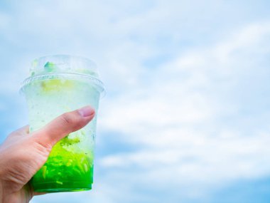 Sol el buzlu holding limon kokteyl limon dilimi üzerinde kopya alanı ile mavi gökyüzü arka plan şeffaf plastik cam. Ekşi yeşil serinletici içecek.