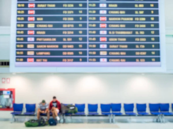 Blurred boarding time monitor screens and passengers sitting on blue chairs in airport hall terminal interior area for arrival passenger.