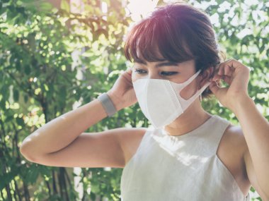 Güzel Asyalı kadın güneş ışığı yeşil yaprakları zemin üzerine beyaz koruyucu N95 toz maske takıyor. Kirliliği kavramı karşı koruma.