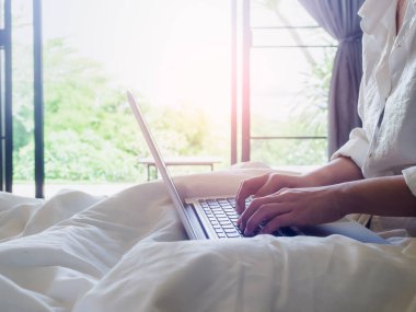 Kadının elleri evin içindeki yatakta dizüstü bilgisayarda yazarken camdan içeri güneş ışığı giriyor. Evde rahatlıkla çalışmak..
