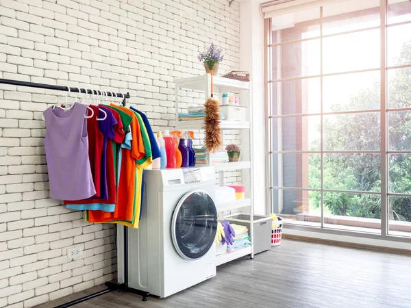 Laundry room interior. Utility room with washing machine, cleaning equipment, home cleaners, clean wipes, hanging colorful shirts on clothesline on white brick wall background near large glass window.