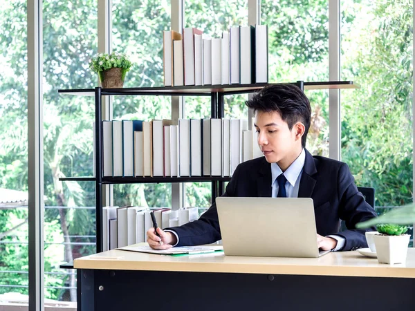 Young Asian businessman in suit working with laptop computer in office with green tree background outside. Man in suit using laptop in well-lit workplace.