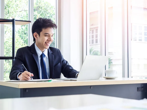 Young smiling Asian businessman in suit working with laptop computer in office. Man in suit using laptop in well-lit workplace.