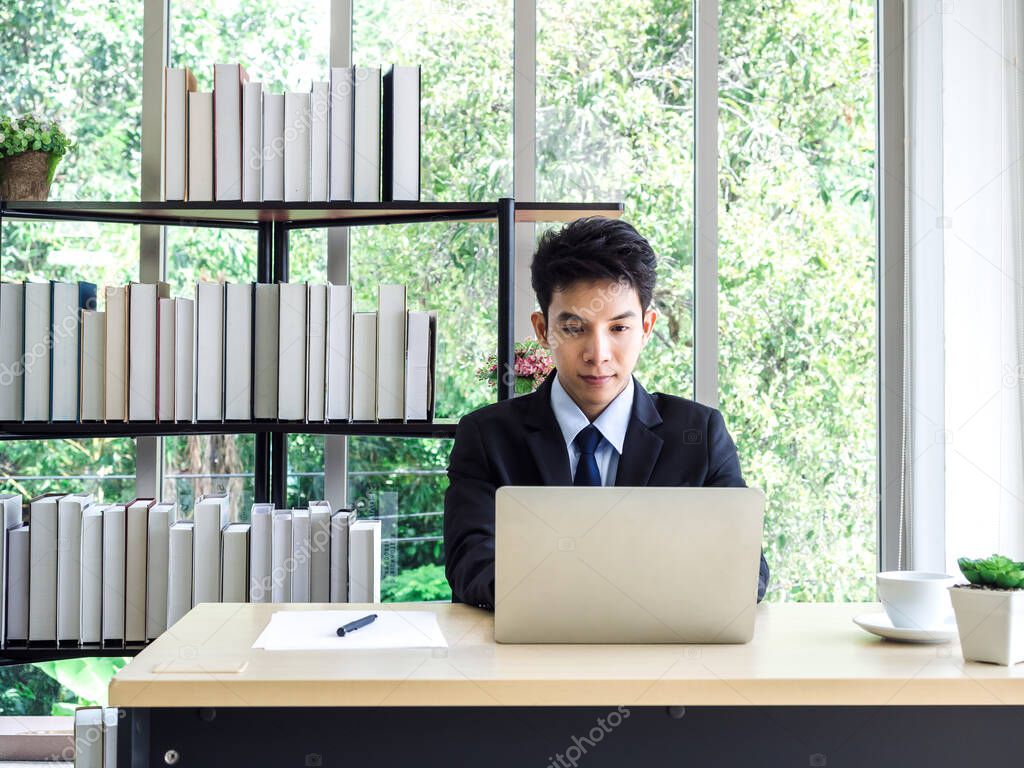 Young Asian businessman in suit working with laptop computer in office with green tree background outside. Man in suit using laptop in well-lit workplace.