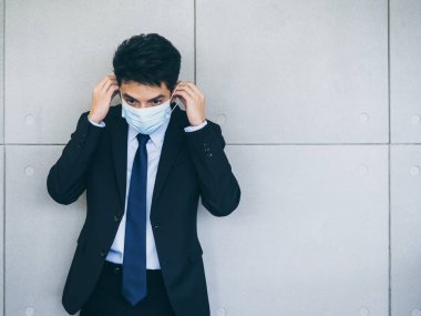 Takım elbiseli genç Asyalı işadamı, ofiste arka planda gri duvar maskesi takıyor. Kopya alanı var. Coronavirüs salgınından sonra yeni normal çalışma tarzı..