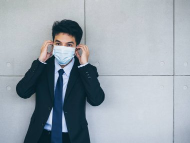 Takım elbiseli genç Asyalı işadamı, ofiste arka planda gri duvar maskesi takıyor. Kopya alanı var. Coronavirüs salgınından sonra yeni normal çalışma tarzı..