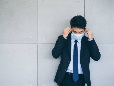 Takım elbiseli genç Asyalı işadamı, ofiste arka planda gri duvar maskesi takıyor. Kopya alanı var. Coronavirüs salgınından sonra yeni normal çalışma tarzı..