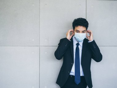 Takım elbiseli genç Asyalı işadamı, ofiste arka planda gri duvar maskesi takıyor. Kopya alanı var. Coronavirüs salgınından sonra yeni normal çalışma tarzı..