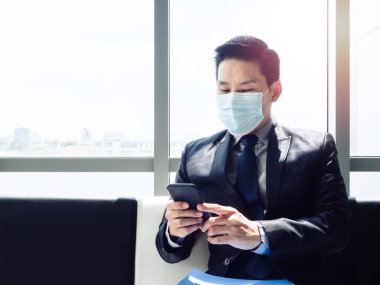Takım elbiseli Asyalı işadamı koruyucu yüz maskesi takıyor ve modern ofis binasında fotokopi alanı olan büyük cam pencerenin yanında otururken akıllı telefon kullanıyor..
