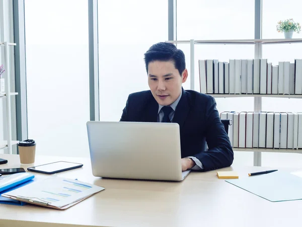 Asian businessman in suit sitting at table and working on laptop computer in office. Manager watching and typing on laptop computer in well-lit workplace. Business, technology and finance concept.