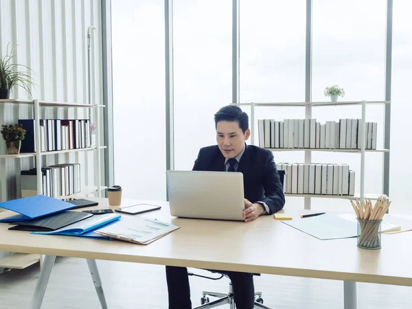 Asian businessman in suit sitting at table and working on laptop computer in office. Manager watching and typing on laptop computer in well-lit workplace. Business, technology and finance concept.