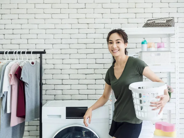 Beautiful young Asian woman housewife standing and holding empty white cloth basket with smiling near washing machine in laundry room.