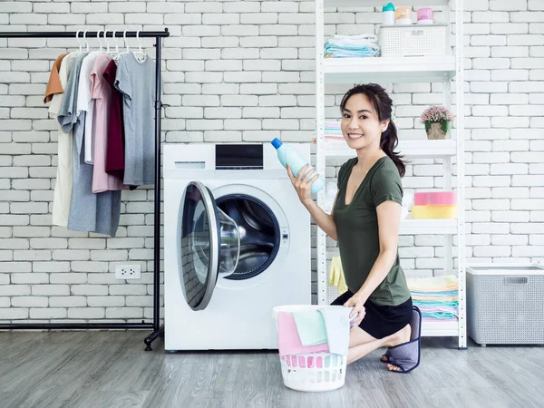 Beautiful young Asian woman housewife sitting and holding liquid laundry detergent, blue bottle with smiling and looking at camera near washing machine in laundry room.