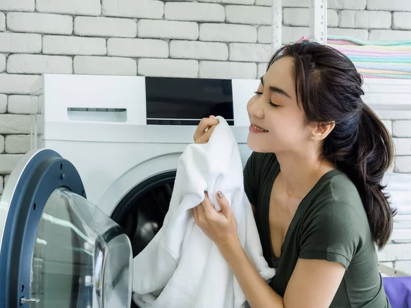 在洗衣房洗衣机里洗完衣服后 年轻美丽的亚洲女性家庭主妇坐在那里 满脸笑容 闻着洁白的毛巾 — 图库照片