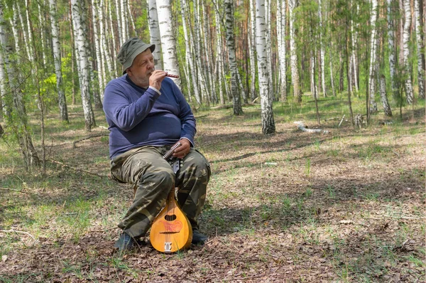 Seniors playing music on woodwind instrument sopilka while sitting on a stool in birch forest