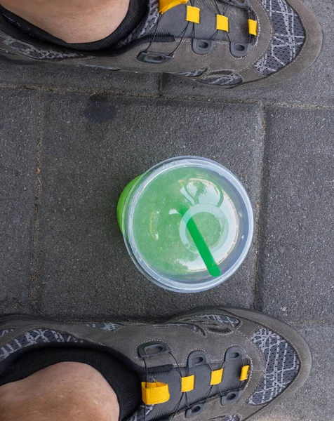 Top view on plastic cup with greenish liquid (mojito) and cocktail tube inside, standing on pavement between human feet cycling shoes