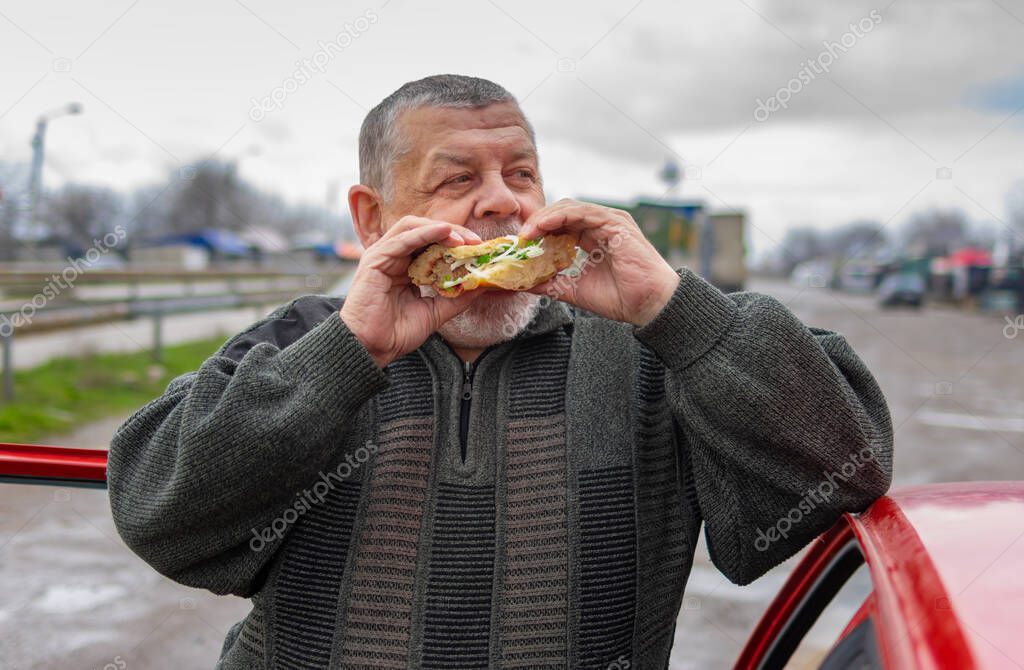 Caucasian senior driver gobbling lyulya kebab in lavash leaning his elbow on the car roo