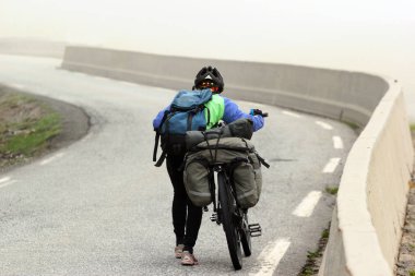 A woman on a bicycle touring trip pushes her bike up the mountain road in foggy weather clipart
