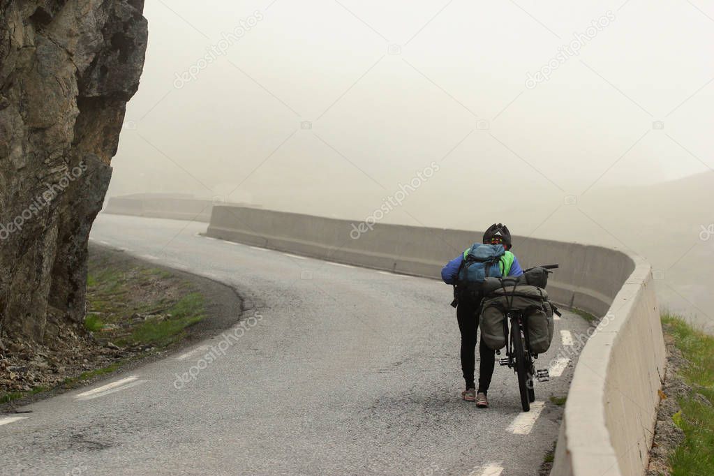 A woman on a bicycle touring trip pushes her bike up the mountain road in foggy weather