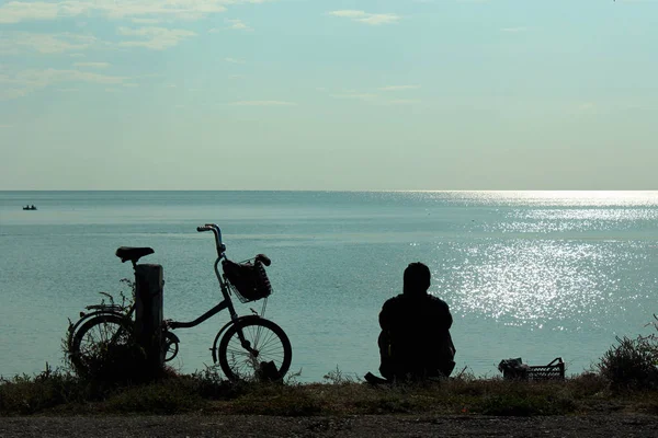 Silhouette di bicicletta e uomo seduto sulla riva del mare e guardando il mare — Foto Stock