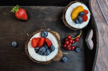 ahşap elemanları ile koyu bir arka plan üzerinde taze hindistan cevizi bir kase renkli taze çilek, yaban mersini ve diğer meyveler
