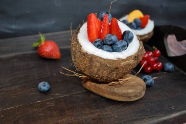 ahşap elemanları ile koyu bir arka plan üzerinde taze hindistan cevizi bir kase renkli taze çilek, yaban mersini ve diğer meyveler