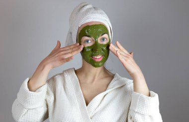 Kafasında havlu olan kadın yeşil kilden kozmetik bir maske yapıyor. Komik ev spa konsepti.