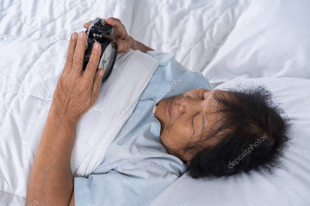 senior woman in bed pressing snooze button on early morning alarm clock 