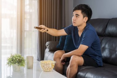 Genç adam elinde uzaktan kumandayla oturma odasında kanepede oturup televizyon seyrediyor. 