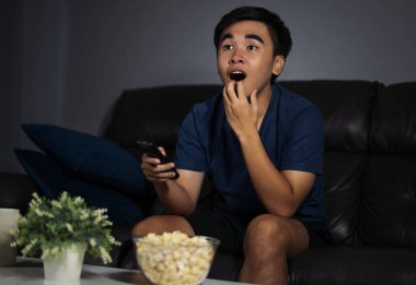 Şok olmuş genç bir adam TV seyrediyor ve geceleri oturma odasında kanepede oturuyor.
