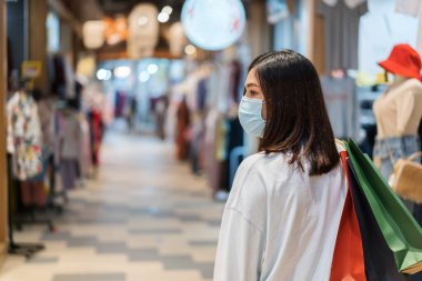 Alışveriş merkezinde alışveriş torbası tutan genç bir kadın ve Coronavirus (Covid-19) salgınını önlemek için tıbbi maske takıyor. yeni normal kavramlar
