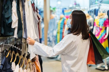 Genç bir kadın alışveriş merkezinde kıyafet seçiyor ve Coronavirus (Covid-19) salgınını önlemek için tıbbi maske takıyor. yeni normal kavramlar