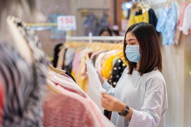 Genç bir kadın alışveriş merkezinde kıyafet seçiyor ve Coronavirus (Covid-19) salgınını önlemek için tıbbi maske takıyor. yeni normal kavramlar