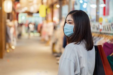 Alışveriş merkezinde çanta taşıyan ve Coronavirus (Covid-19) salgınını önlemek için tıbbi maske takan genç bir kadın. yeni normal kavramlar