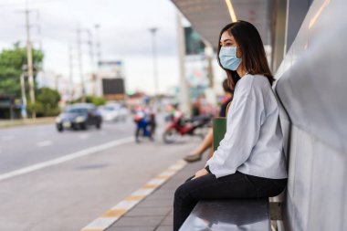 Otobüs durağında otobüs bekleyen ve Coronavirus (covid-19) salgınının yayılması için maske takan genç bir kadın.