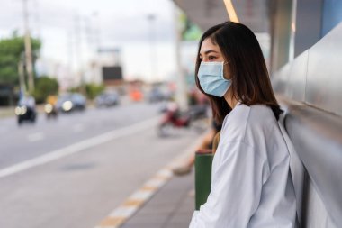Otobüs durağında otobüs bekleyen ve Coronavirus (covid-19) salgınının yayılması için maske takan genç bir kadın.