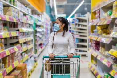 Markette alışveriş arabalı genç bir Asyalı kadın ve Coronavirus (covid-19) salgınını önlemek için tıbbi maske takıyor. yeni normal kavram