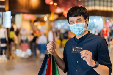 Coronavirus (Covid-19) salgınını önlemek için tıbbi maske takan genç Asyalı bir adam ve alışveriş merkezinde kredi kartı tutuyor. yeni normal kavramlar