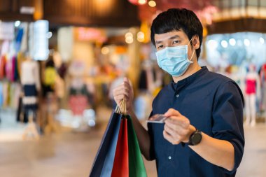 Coronavirus (Covid-19) salgınını önlemek için tıbbi maske takan genç Asyalı bir adam ve alışveriş merkezinde kredi kartı tutuyor. yeni normal kavramlar