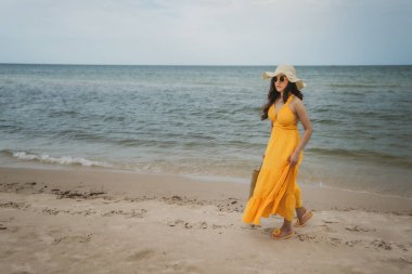 Sarı elbiseli genç bir kadın deniz sahilinde yürüyor.