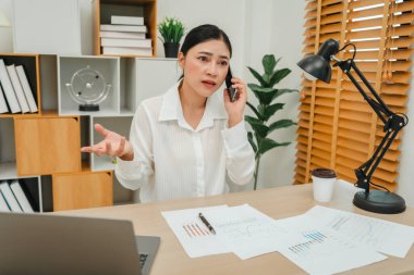 Stresli genç bir kadın akıllı telefonuyla konuşuyor. Ofiste belge ve dizüstü bilgisayarla çalışmak
