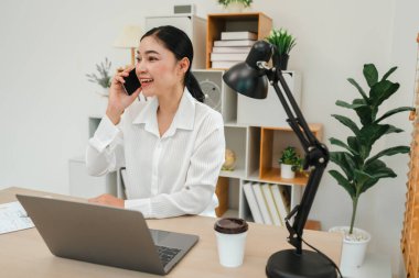 Ofiste dizüstü bilgisayarla çalışırken akıllı telefonla konuşan mutlu genç bir kadın.
