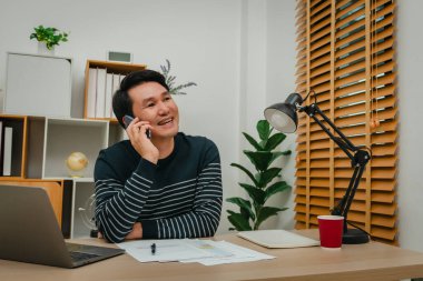 Ofiste doküman ve dizüstü bilgisayarla çalışırken akıllı telefonla konuşan mutlu genç adam.