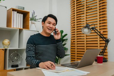 Ofiste bilgisayarla çalışırken akıllı telefonla konuşan mutlu bir adam.