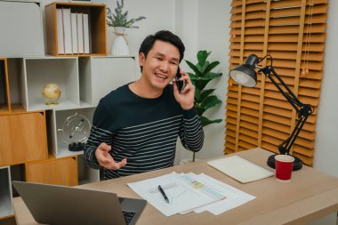 Ofiste doküman ve dizüstü bilgisayarla çalışırken akıllı telefonla konuşan mutlu genç adam.